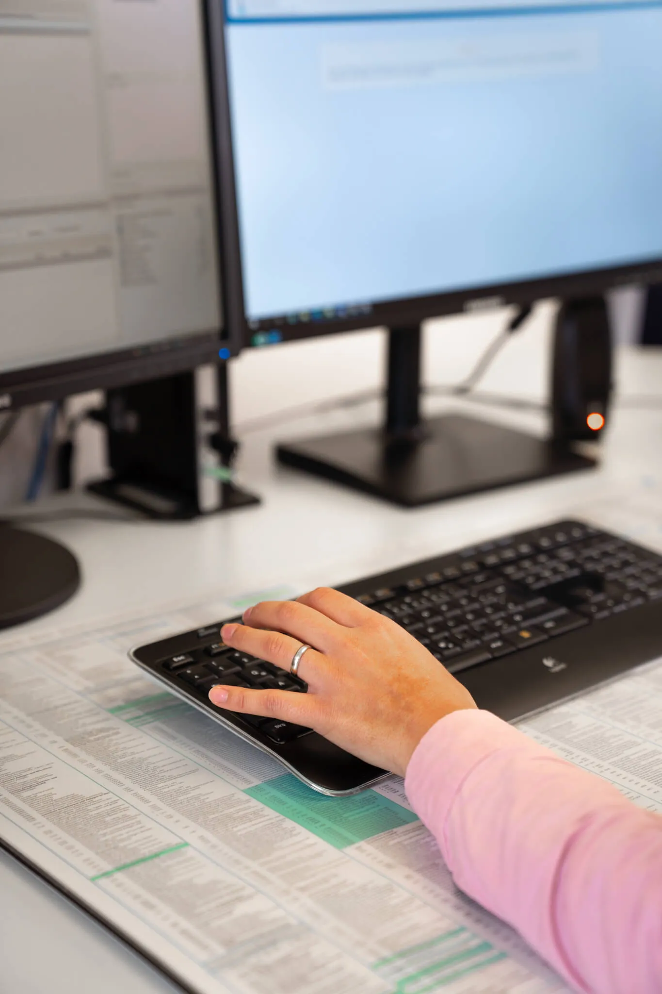 Nahaufnahme einer Hand mit einem Ring, die auf einer Computertastatur liegt. Im Hintergrund zwei Monitore mit geöffneten Programmen.