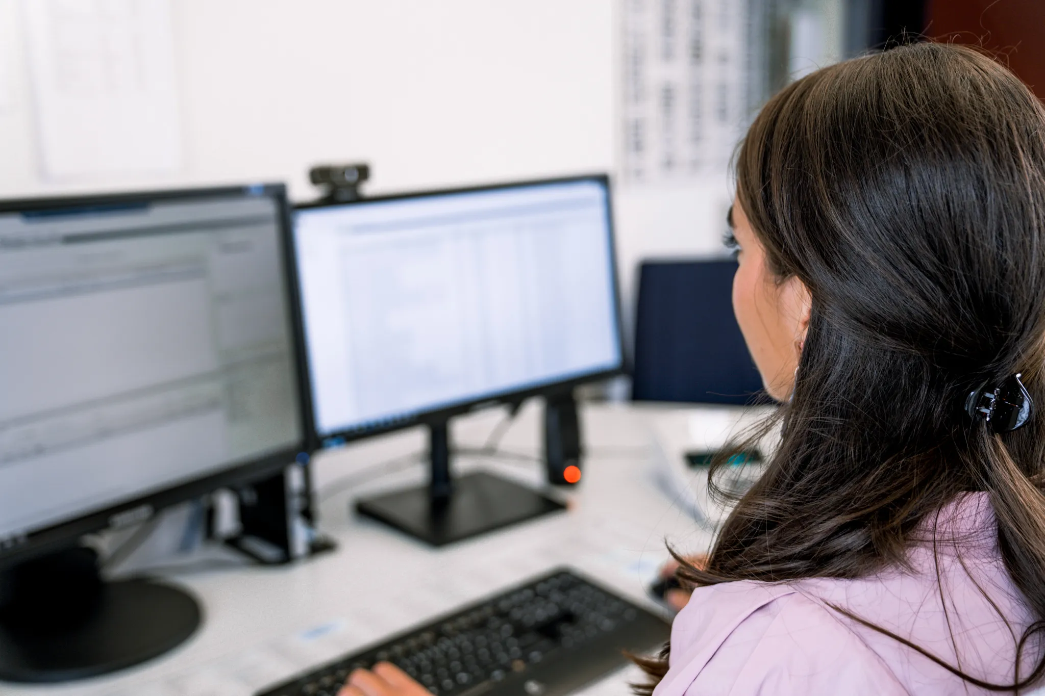 Frau mit dunklen Haaren von hinten, arbeitet an zwei Monitoren im Büro.