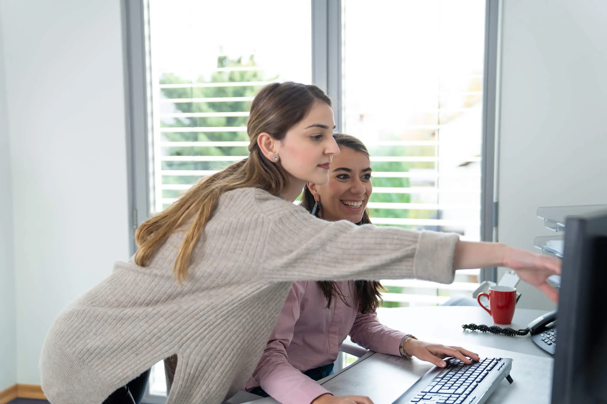 Zwei junge Frauen arbeiten gemeinsam an einem Computer. Eine sitzt und lächelt, während die andere steht und auf den Monitor zeigt.
