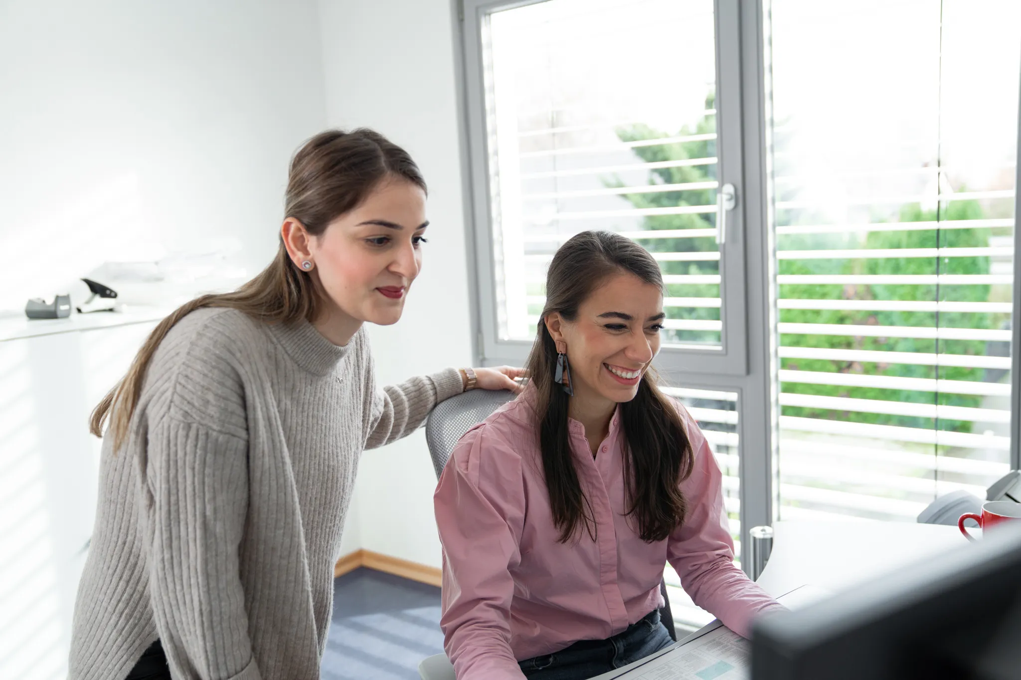 Zwei junge Frauen arbeiten gemeinsam an einem Computer; eine sitzt lachend am Schreibtisch, die andere lehnt sich von der Seite heran.