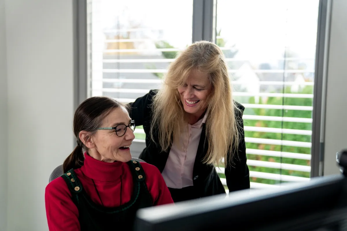 Zwei Mitarbeiterinnen unterhalten sich am Arbeitsplatz vor einem Computerbildschirm.