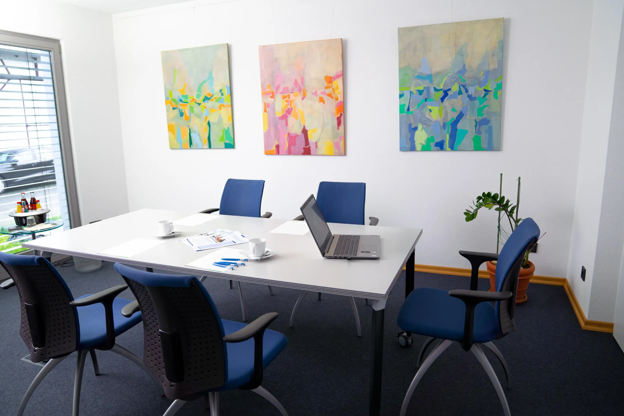 Moderner Besprechungsraum mit vier blauen Stühlen um einen weißen Tisch, auf dem ein Laptop, Tassen und Schreibutensilien liegen. Bunte Bilder hängen an der Wand.