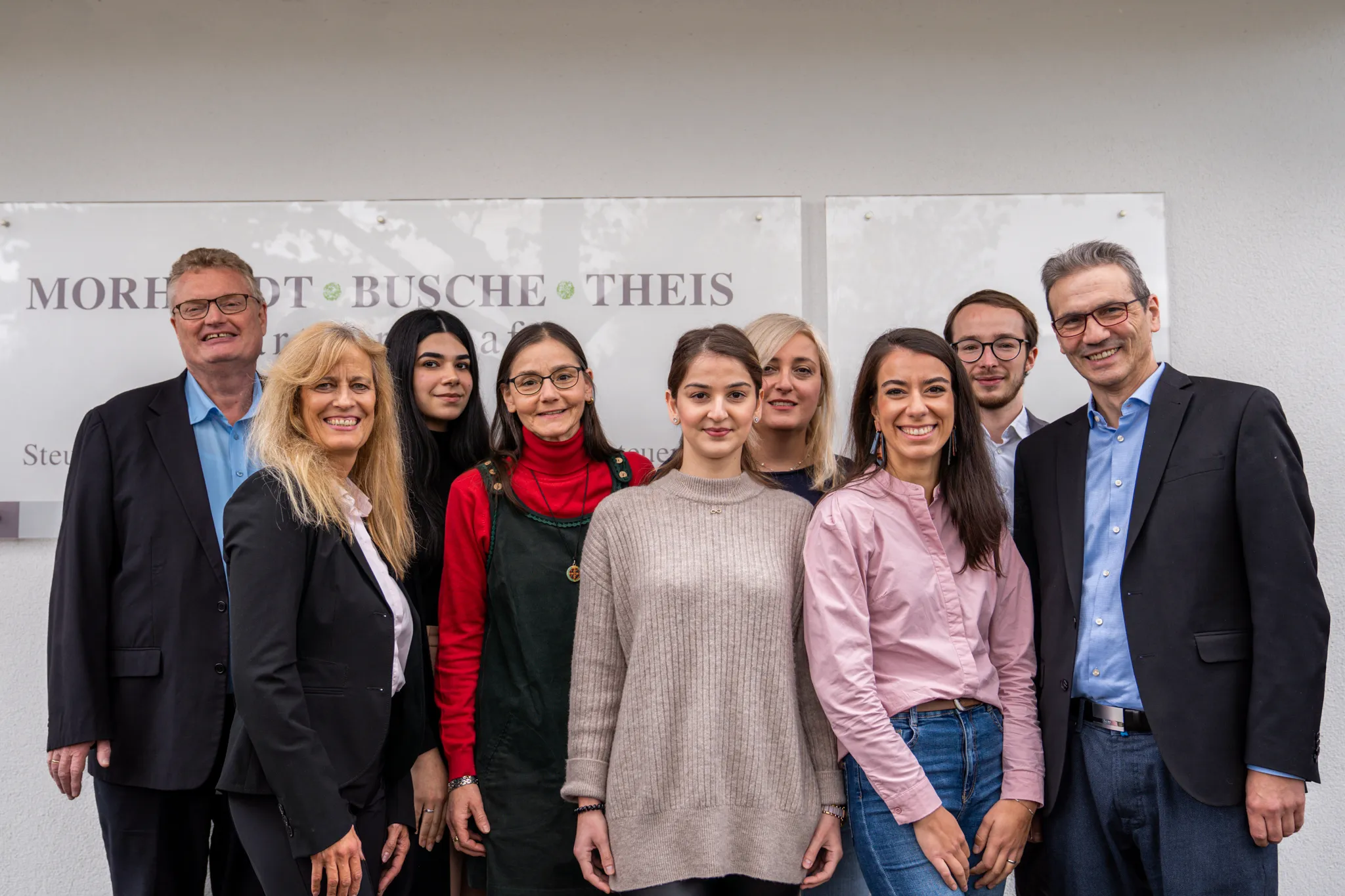 Gruppenfoto von neun Kolleginnen und Kollegen vor einem Firmenschild mit dem Schriftzug ‚Morkhof Busche Theis‘. Alle lächeln in die Kamera.