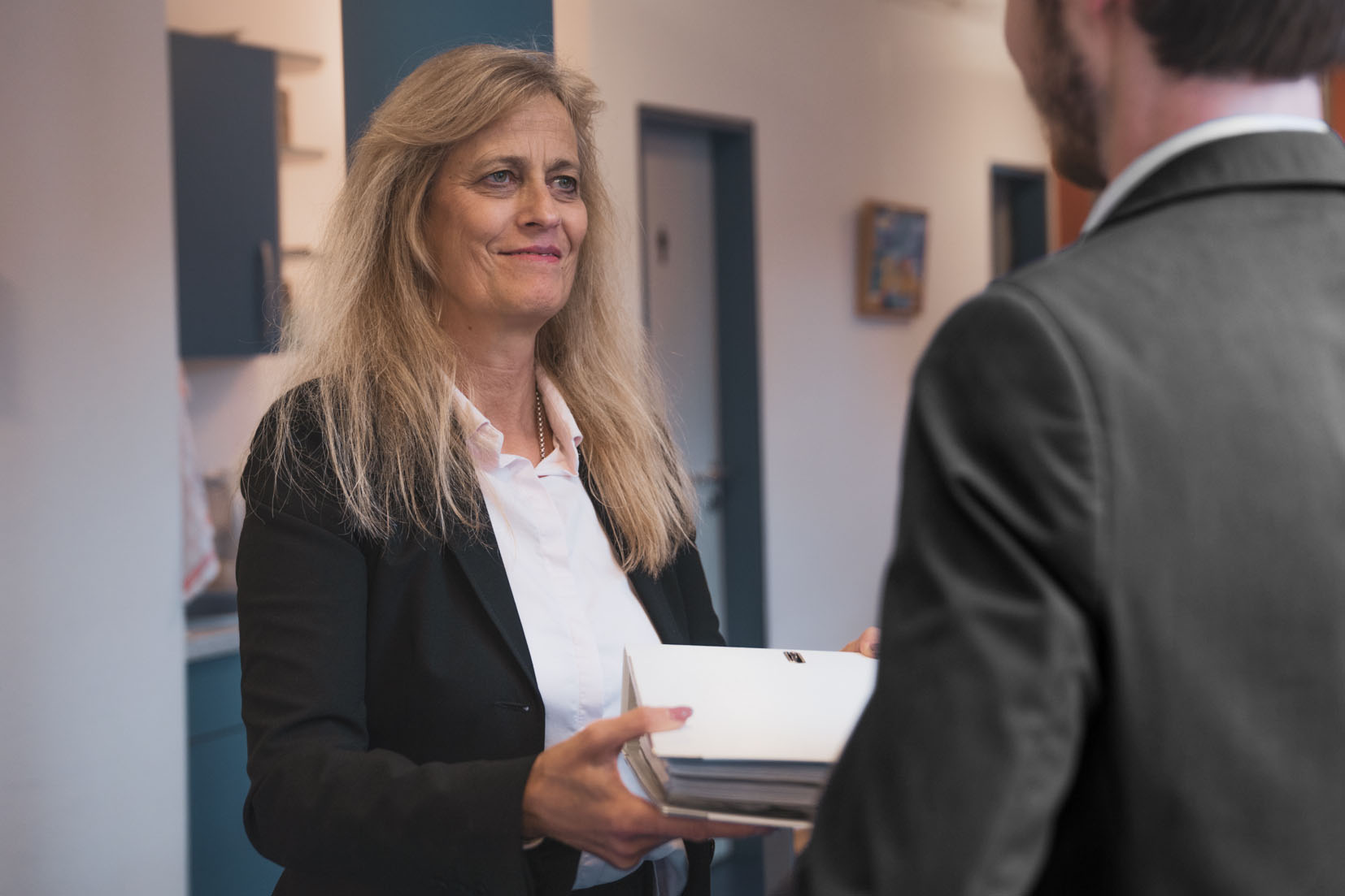 Eine Frau mit langen blonden Haaren übergibt einem Mann einen Stapel Aktenordner. Beide tragen Business-Kleidung.