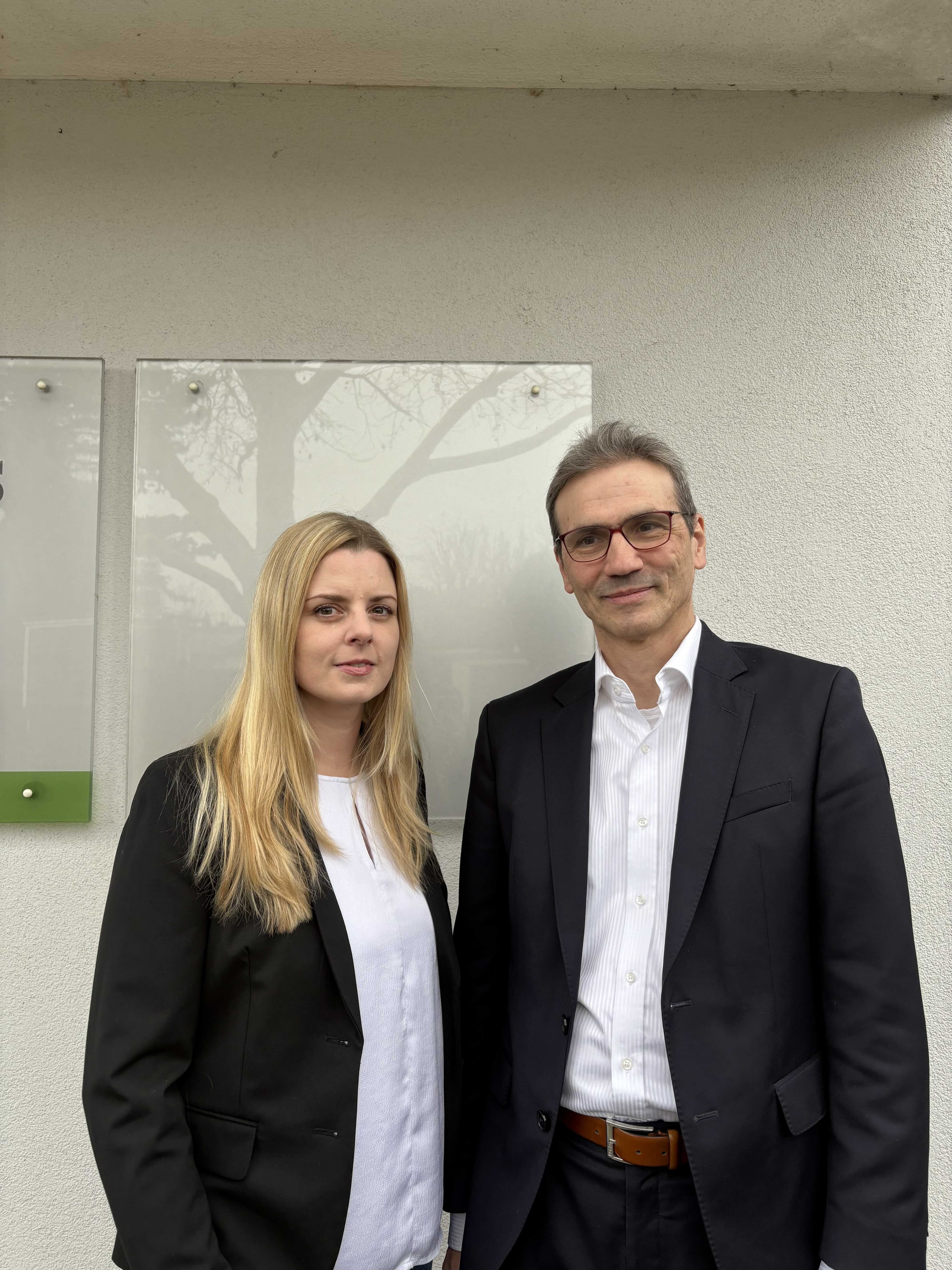 Eine Frau mit langen blonden Haaren und ein Mann mit grauen Haaren und Brille stehen nebeneinander vor einer weißen Wand mit Glas-Schildern. Beide tragen Business-Kleidung.